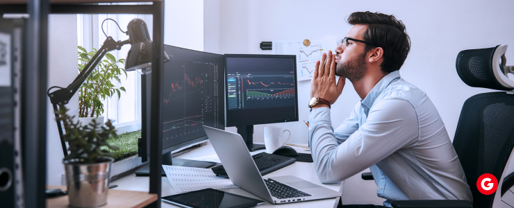 A man seated at a desk with dual monitors and a laptop, engaged in short-duration forex trading.